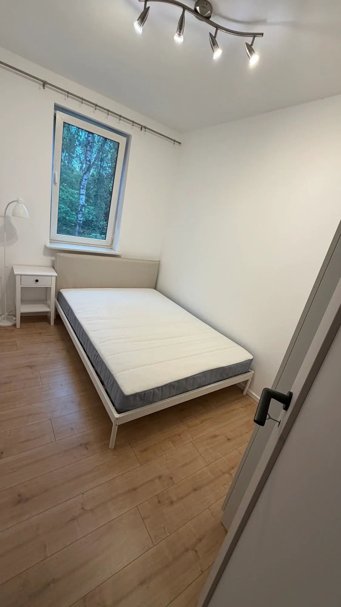 Złożona rama łóżka z materacem w pustym pokoju, obok szafka nocna i lampa